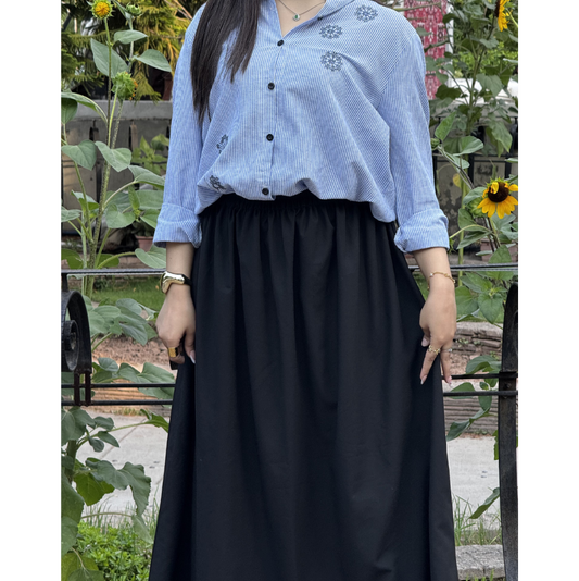 A person wearing a light blue and white vertically striped button-up shirt with small floral embroidery on the chest, paired with a long black skirt, standing outdoors in front of a garden with sunflowers.