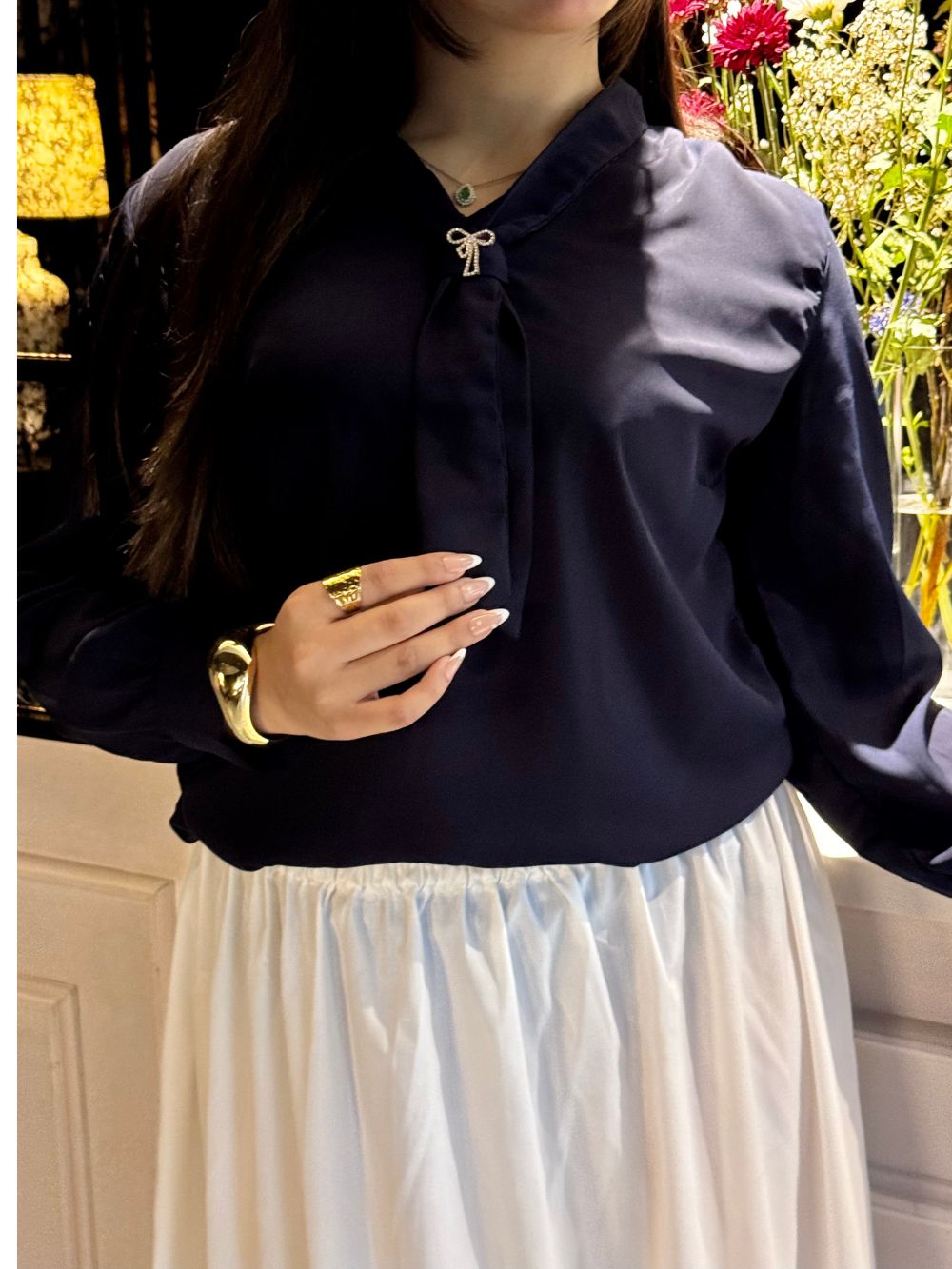 A woman wearing a dark navy long-sleeve blouse with a bow-shaped collar detail and a white skirt, shown in a styled indoor setting with warm lighting and flowers in the background.