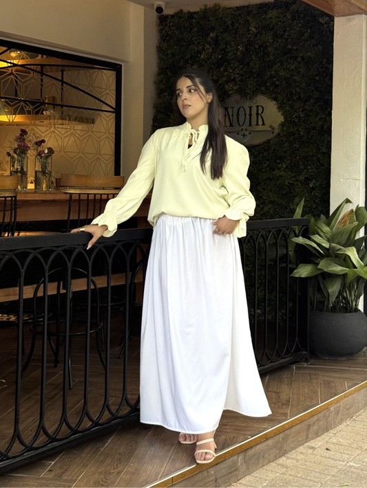 A woman wearing a pale yellow long-sleeve blouse with a tie-neck detail and a long white skirt, standing on a balcony with a black railing in front of a green wall.