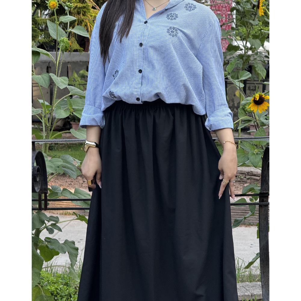 A person wearing a light blue and white vertically striped button-up shirt with small floral embroidery on the chest, paired with a long black skirt, standing outdoors in front of a garden with sunflowers.