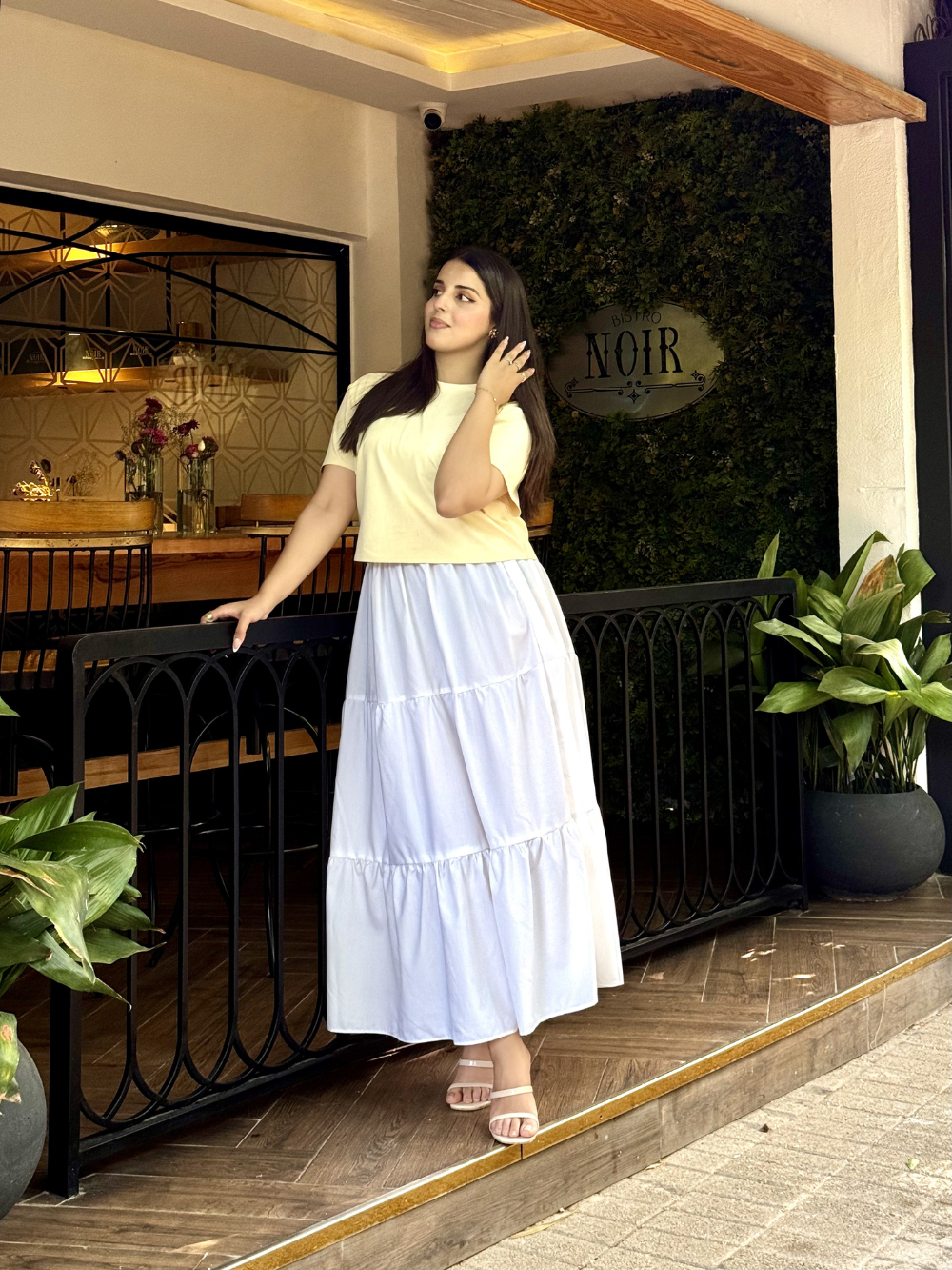 A woman stands on a restaurant porch wearing a pale yellow short-sleeve top and a long white tiered skirt. The setting includes a black railing, potted plants, a wood floor, and a greenery wall with a sign reading NOIR.