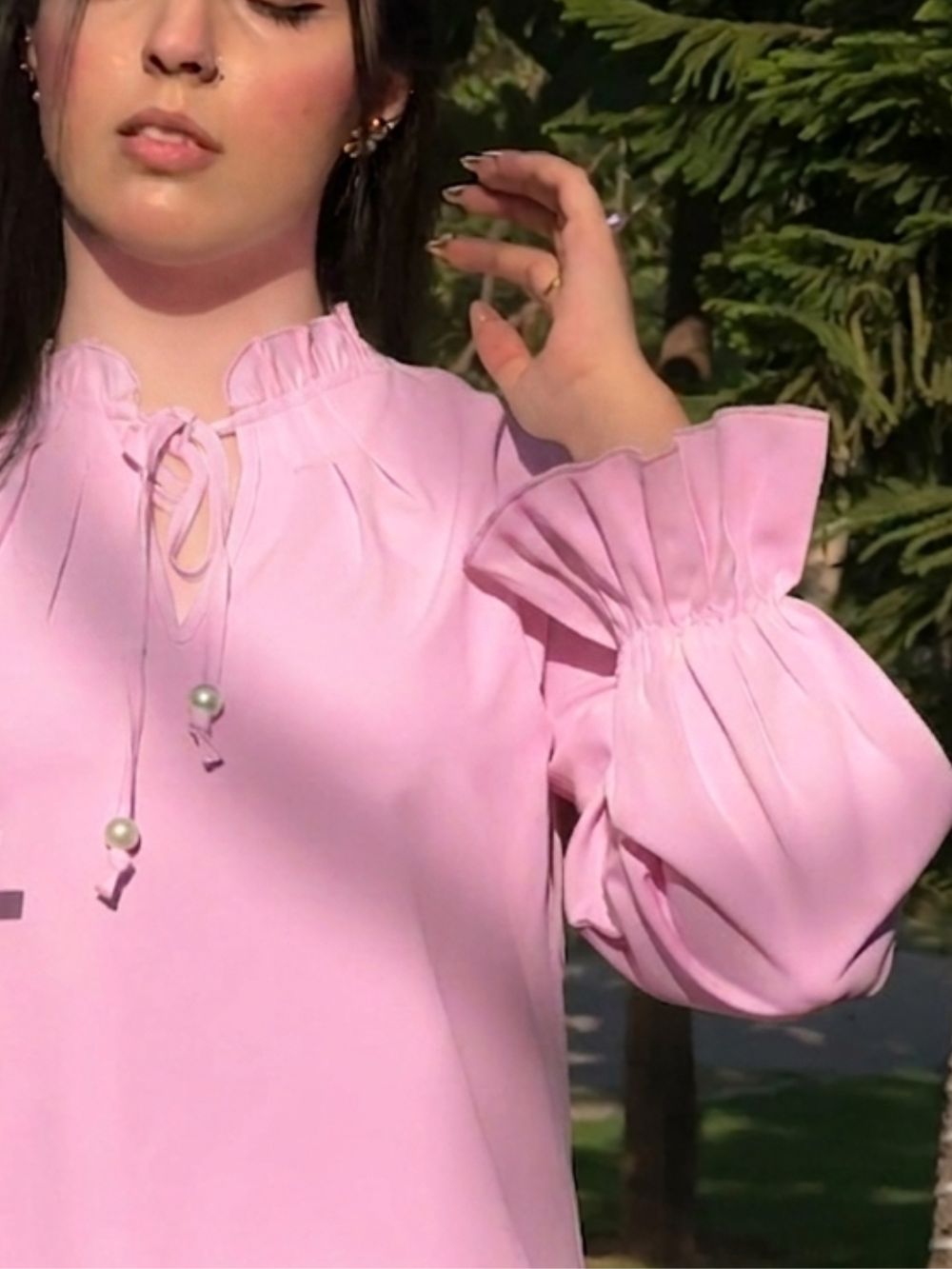 A close-up of a woman wearing a light pink blouse with a ruffled high neck, front tie detail, and voluminous gathered sleeves, photographed outdoors with green foliage in the background.